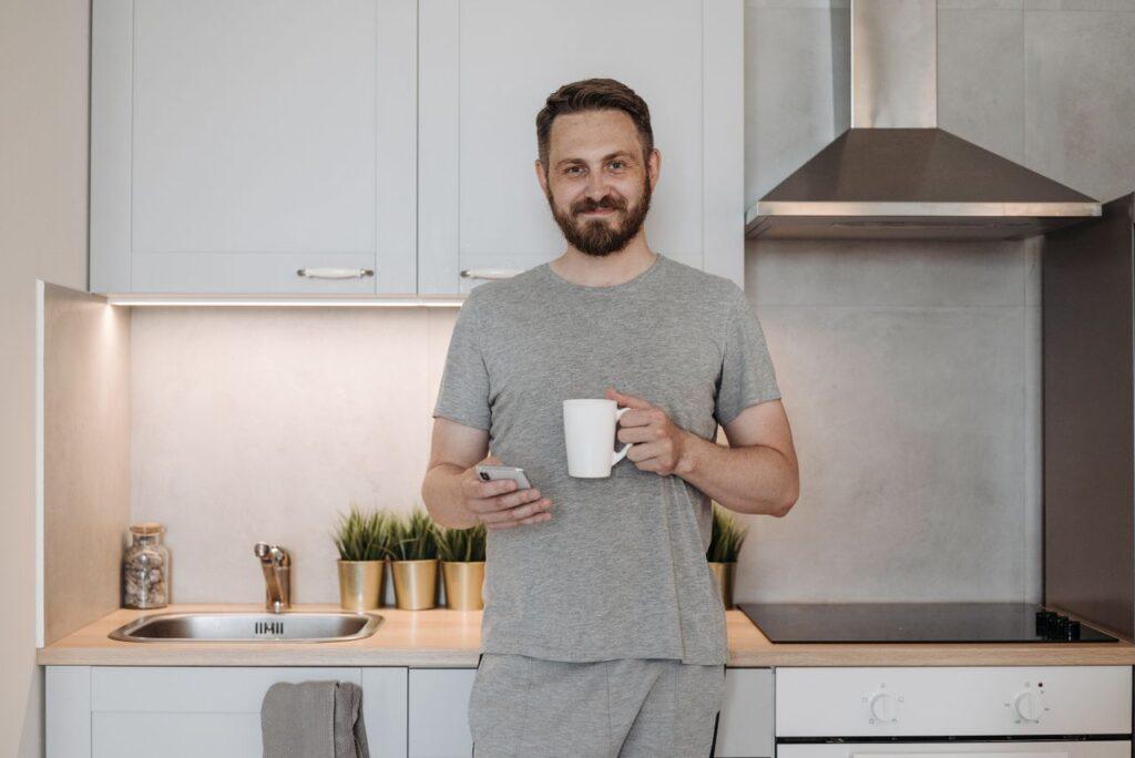 Ein Mann steht morgens entspannt in der Küche mit einer Tasse Kaffee – Sinnbild für einen ganz normalen Dienstag im Alltag